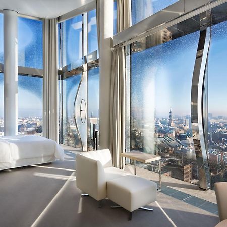 The Westin Hamburg Elbphilharmonie Hotell Eksteriør bilde The photo shows a modern hotel room with a stunning, panoramic view through large, uniquely designed windows. The room features a sleek, minimalist aesthetic with light-colored furnishings, including a white bed and a couple of chairs. The walls are 