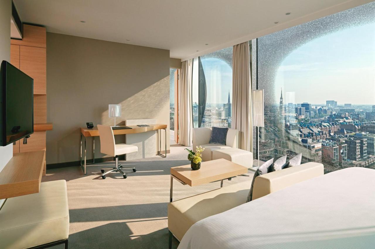 The Westin Hamburg Elbphilharmonie Hotell Eksteriør bilde The photo depicts a modern hotel room featuring a contemporary design. The space includes a large window that offers a panoramic view of the city beyond. Inside, there are stylish furniture elements such as a small dining table and chairs, a desk wit