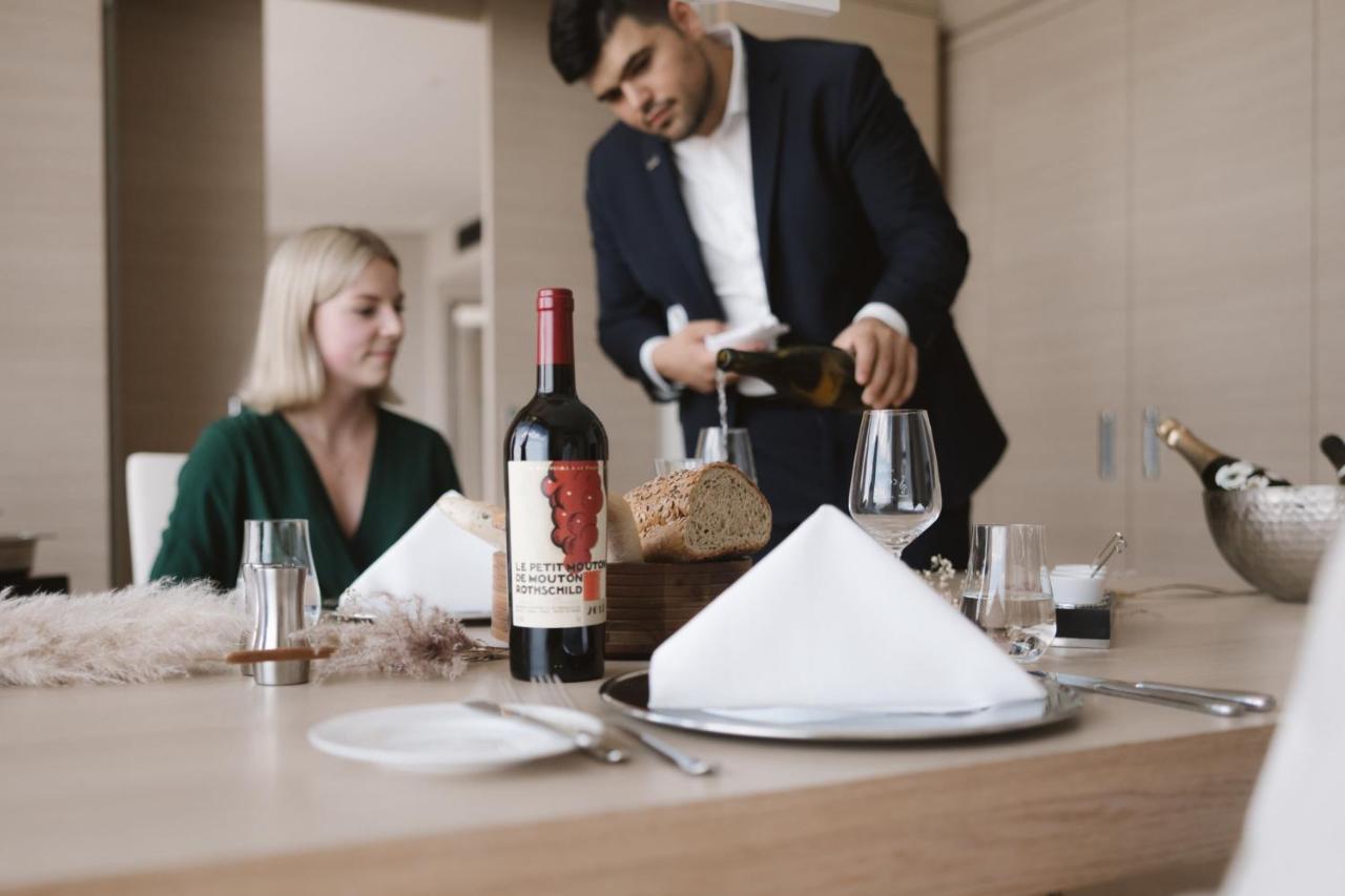The Westin Hamburg Elbphilharmonie Hotell Eksteriør bilde The photo shows a dining scene featuring a well-dressed man pouring wine into a glass while a woman sits at the table. The table is elegantly set with a folded napkin, wine glasses, and a bottle of red wine placed prominently. There is also a loaf of