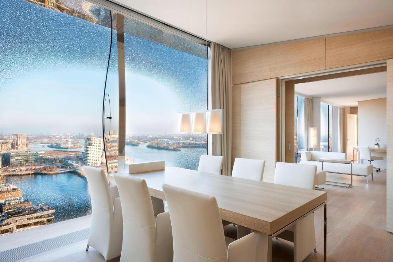 The Westin Hamburg Elbphilharmonie Hotell Eksteriør bilde The photo shows a modern dining area with a large, light-colored table surrounded by white chairs. The space features a panoramic window that offers a stunning view of a body of water and a cityscape. The decor is minimalistic and elegant, with smoot