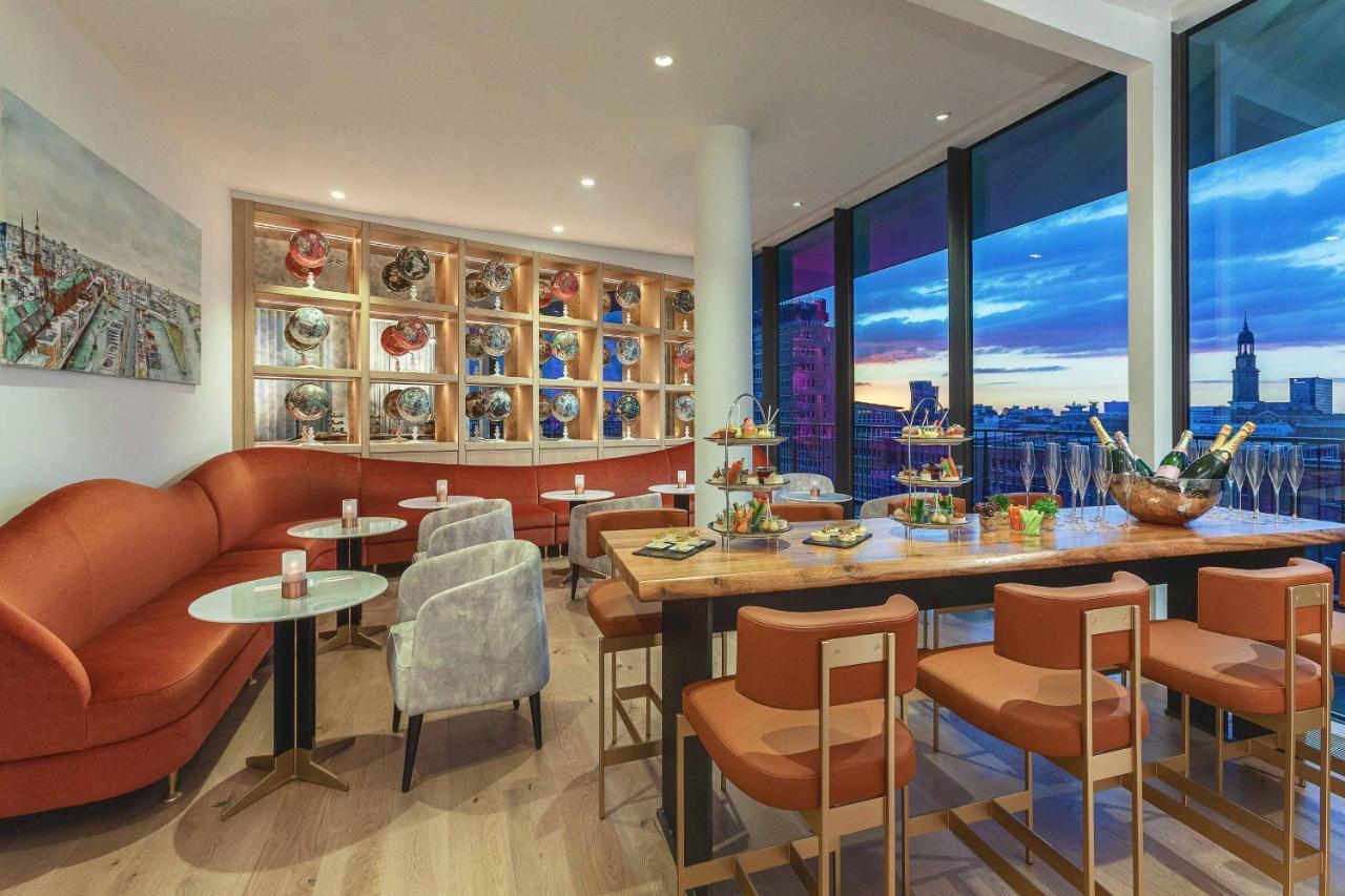 The Westin Hamburg Elbphilharmonie Hotell Eksteriør bilde The photo shows a modern and stylish restaurant or café interior. It features a combination of comfortable seating, including a curved, orange sofa and gray chairs. The tables are round with light-colored tops, and there are several tiered stands dis