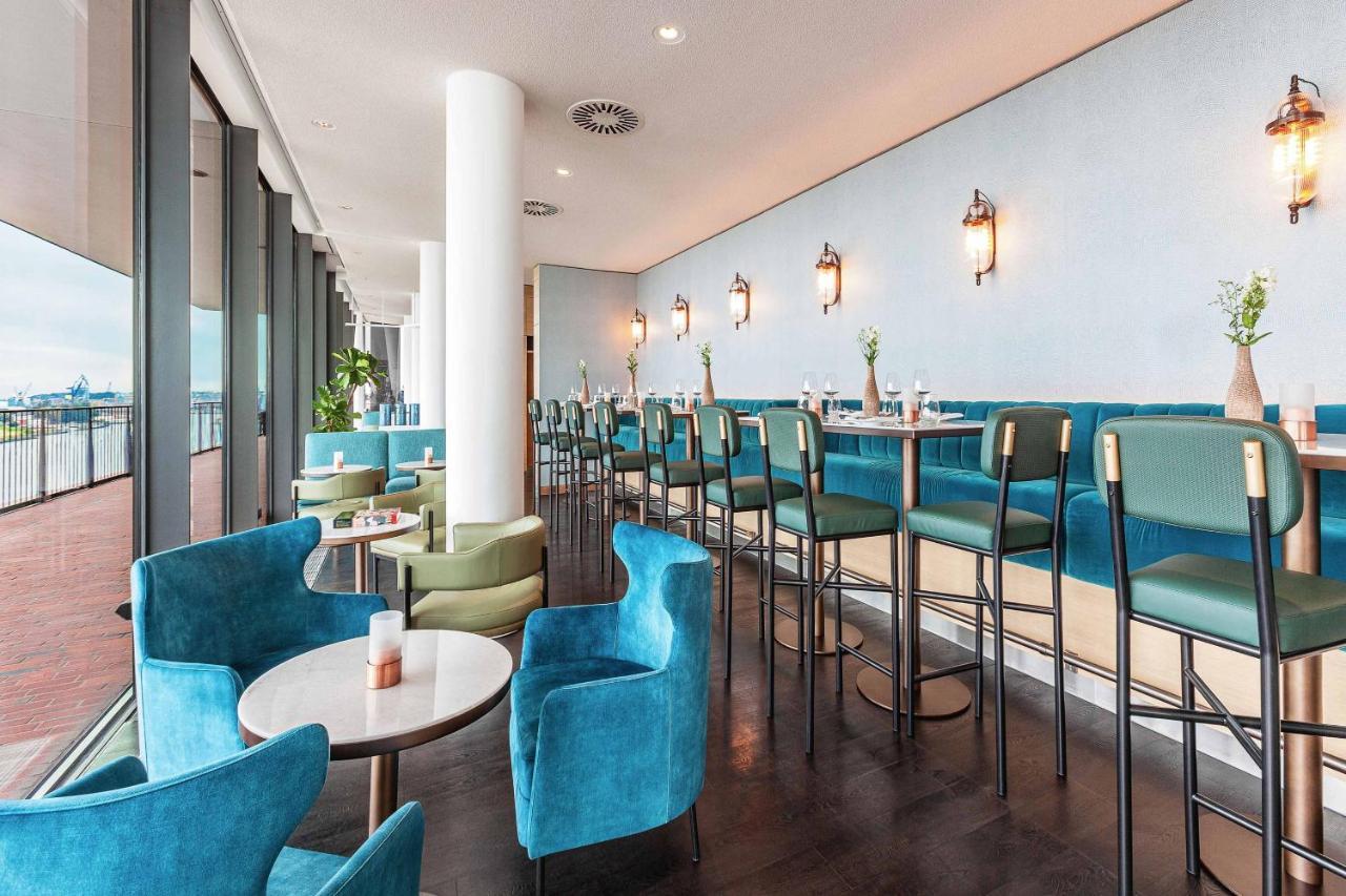 The Westin Hamburg Elbphilharmonie Hotell Eksteriør bilde The photo shows a modern and stylish restaurant or café interior. It features a long row of high-backed bar stools at a counter, complemented by plush seating in the form of rounded chairs and small tables arranged throughout the space. The color sch