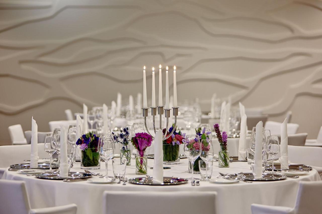 The Westin Hamburg Elbphilharmonie Hotell Eksteriør bilde The photo depicts an elegantly set round dining table. In the center, there is a candelabra with several tall candles, providing a soft, warm light. Surrounding the candelabra are small glass vases filled with colorful flowers, adding a vibrant touch