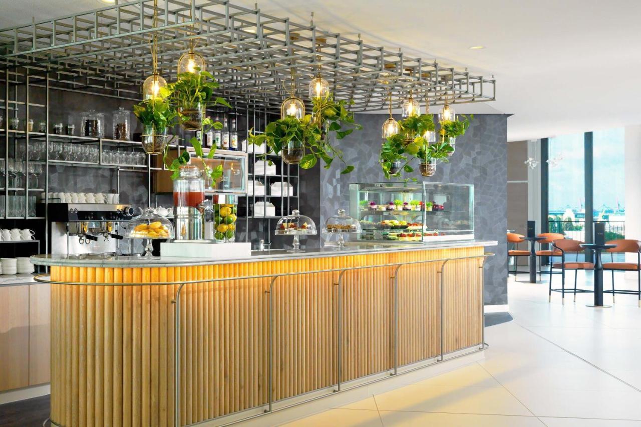 The Westin Hamburg Elbphilharmonie Hotell Eksteriør bilde The photo shows a modern café or bar setting with a sleek wooden counter that features vertical slats. Above the counter, there is a grid-like framework hanging from the ceiling, adorned with various green plants that add a touch of nature to the int