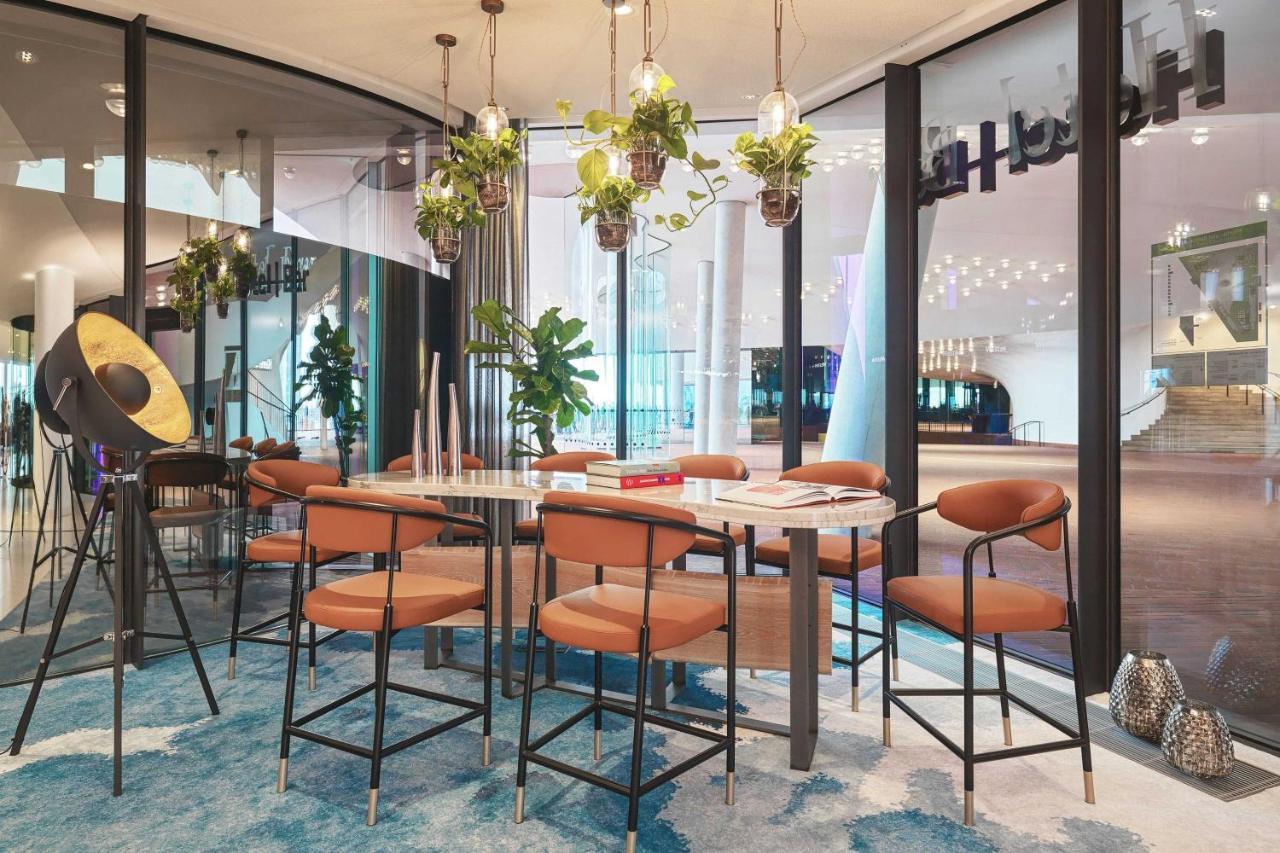 The Westin Hamburg Elbphilharmonie Hotell Eksteriør bilde The image depicts a modern indoor seating area designed for gatherings or meetings. There is a long table surrounded by several bar-style chairs with brown upholstery. Above the table, there are hanging plants that add a touch of greenery to the spac