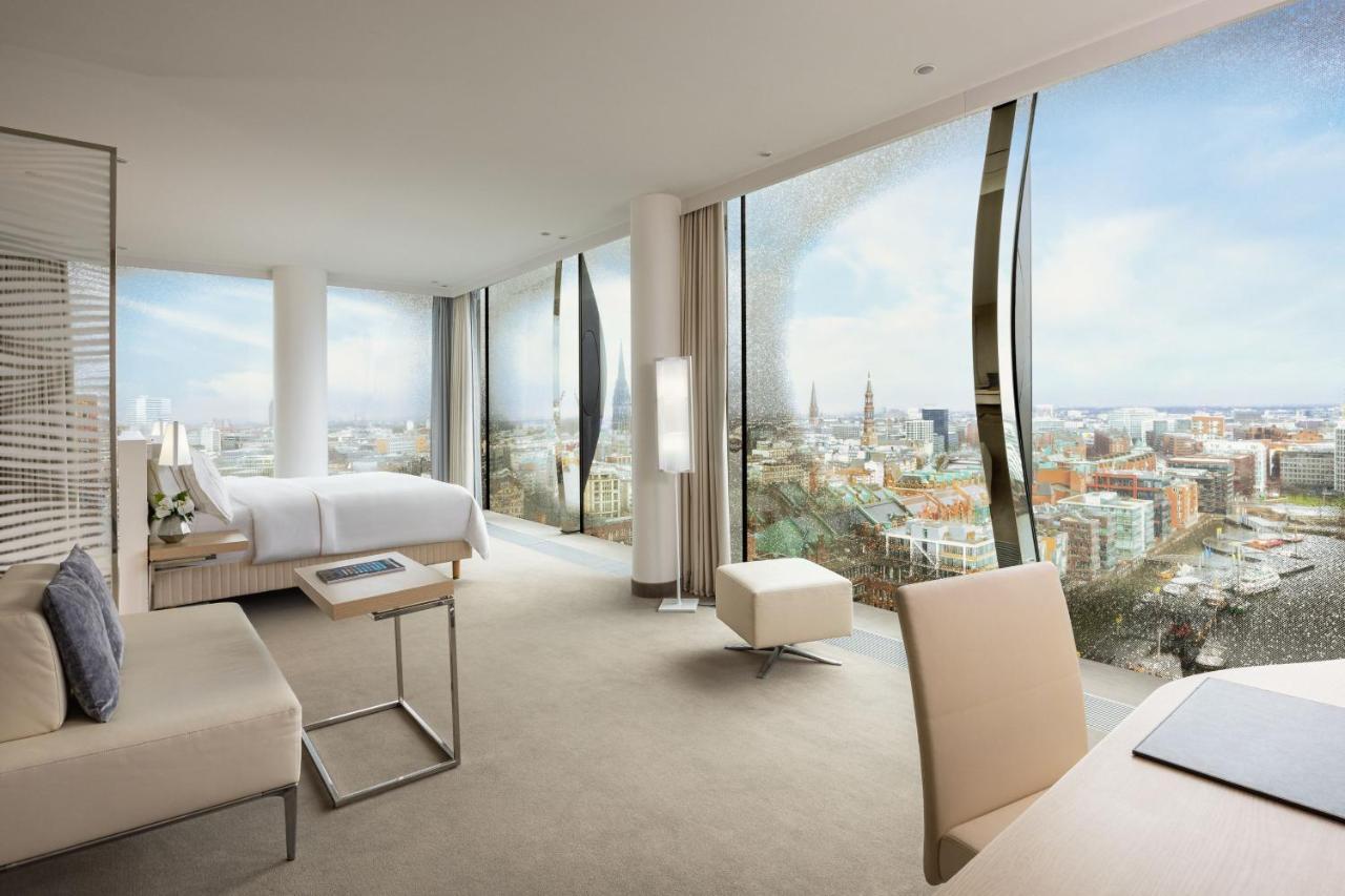 The Westin Hamburg Elbphilharmonie Hotell Eksteriør bilde The photo shows a modern hotel room with large floor-to-ceiling windows that provide a panoramic view of a city landscape. The room features a minimalist design with neutral tones. There's a king-sized bed positioned in the center, a sleek desk and c