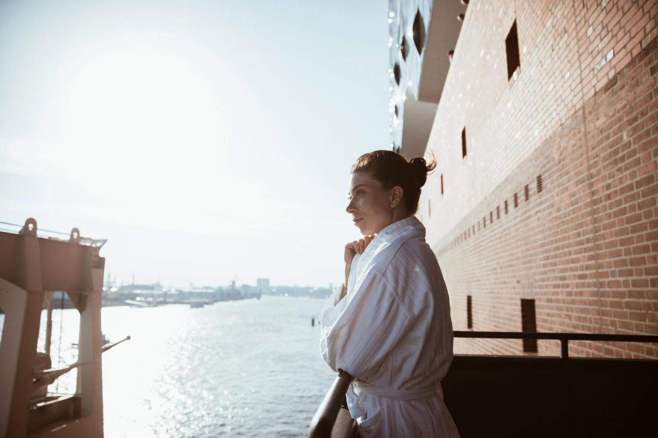 The Westin Hamburg Elbphilharmonie Hotell Eksteriør bilde The photo depicts a person standing on a balcony, looking out over a body of water. The individual is wearing a white bathrobe, and their hair is styled in a bun. The background features a scenic view with sunlight illuminating the scene, creating a 