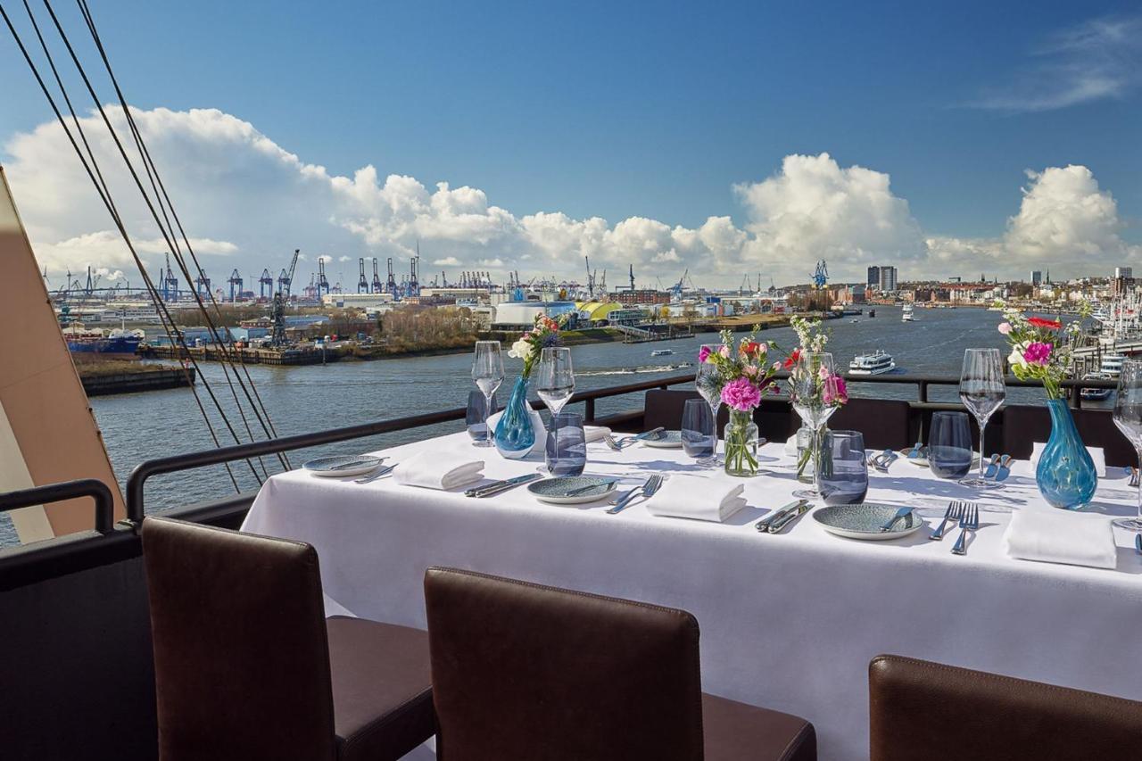 The Westin Hamburg Elbphilharmonie Hotell Eksteriør bilde The photo shows a beautifully set dining table on a rooftop or terrace with a view of a body of water. The table is adorned with a white tablecloth, elegant tableware, and multiple vases filled with colorful flowers. In the background, there are boat