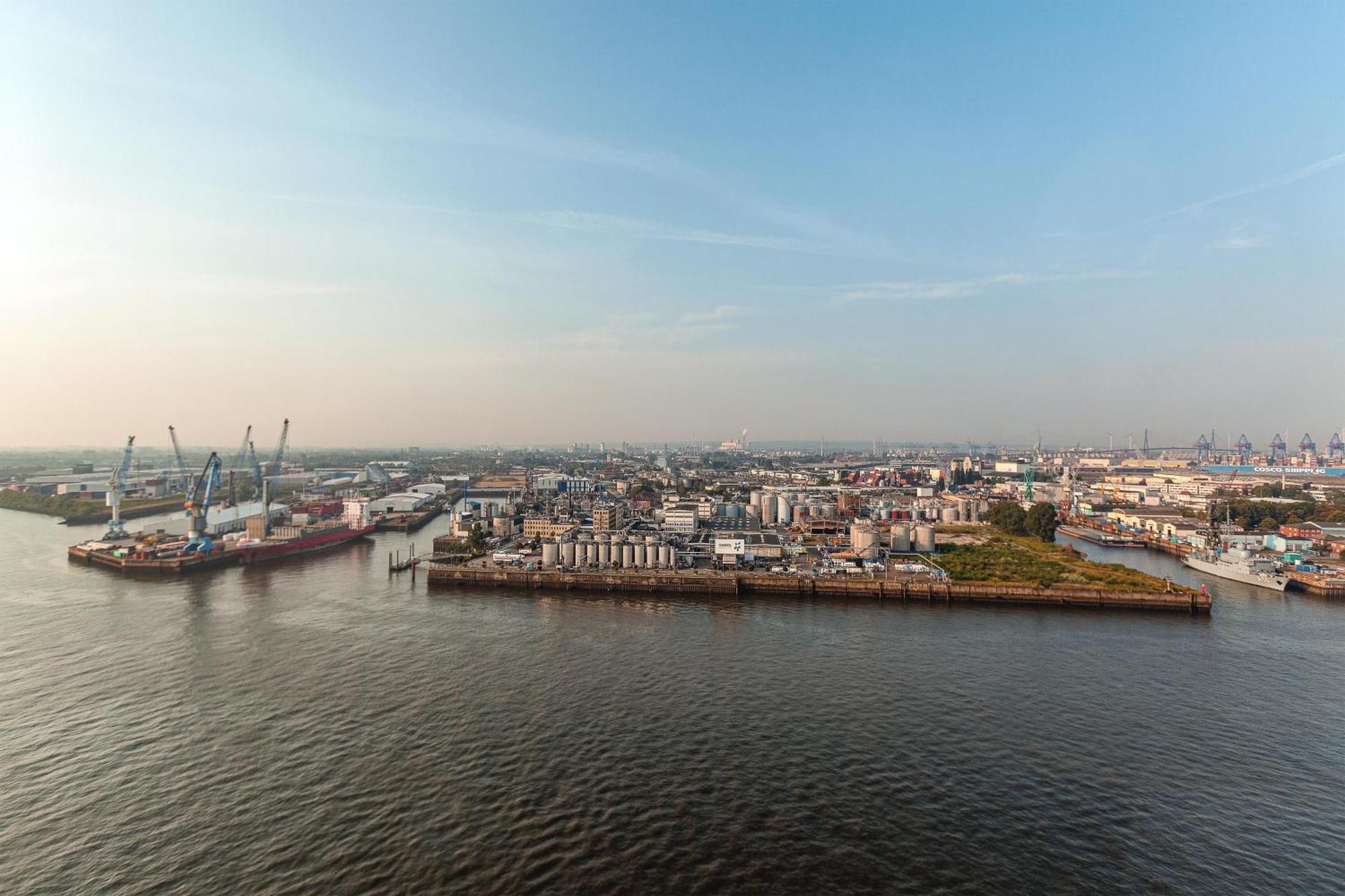 The Westin Hamburg Elbphilharmonie Hotell Eksteriør bilde The photo shows a coastal area with a large body of water in the foreground. To the left, there are industrial structures like cranes and cargo ships, indicating an active port or shipping area. In the background, there are various buildings, which a