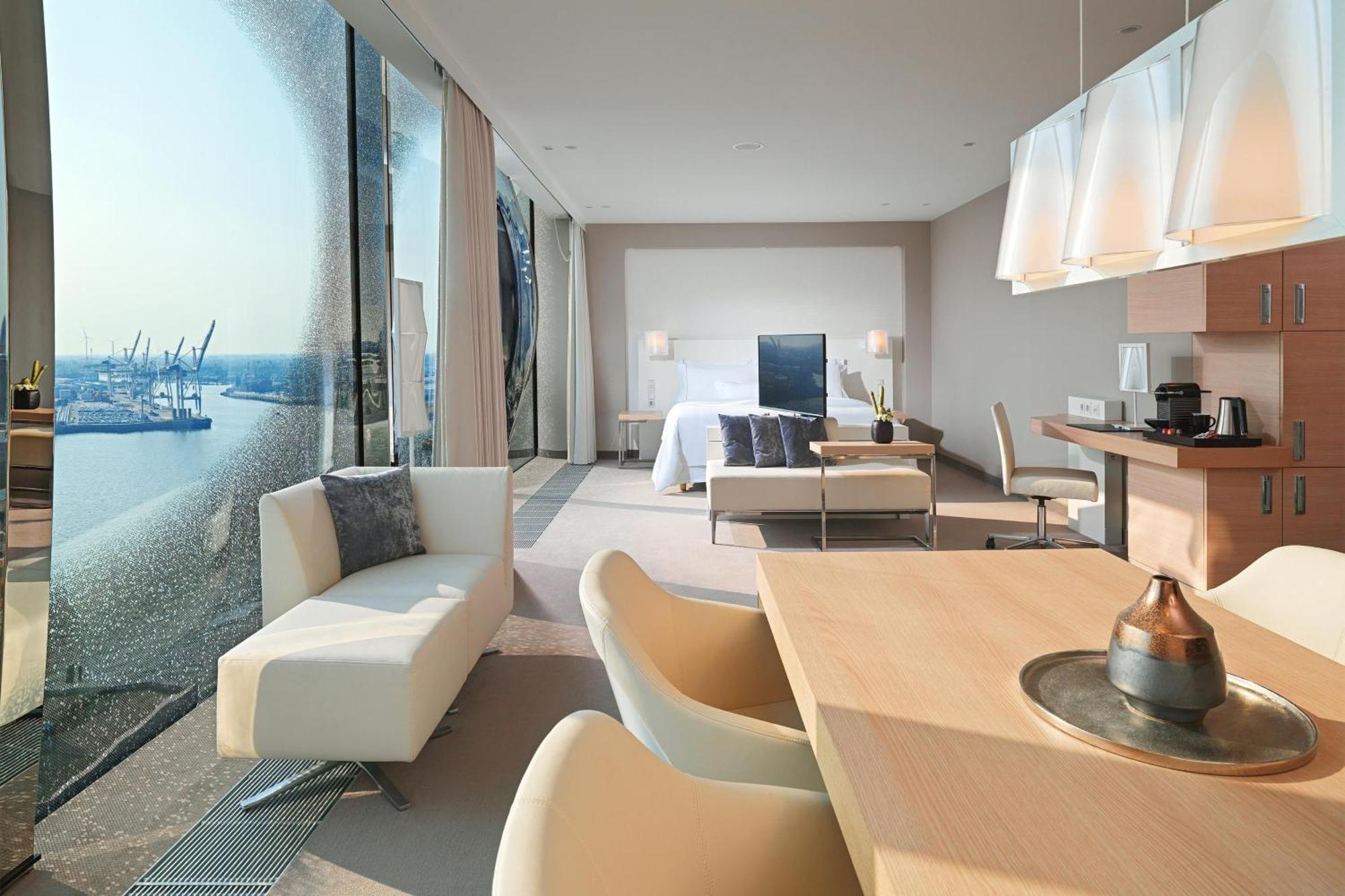 The Westin Hamburg Elbphilharmonie Hotell Eksteriør bilde The photo shows a modern hotel room with a spacious layout. On one side, there are large floor-to-ceiling windows that offer a view of the outside, likely showcasing a waterfront or cityscape. Inside, there's a bed with a minimalist design, accompani
