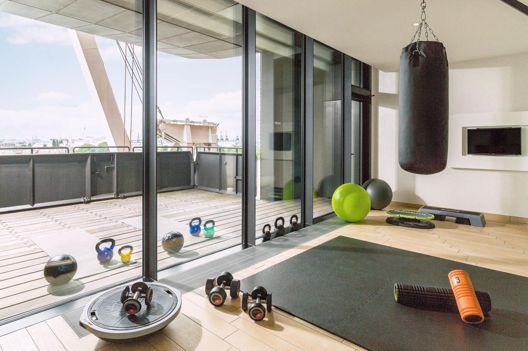The Westin Hamburg Elbphilharmonie Hotell Eksteriør bilde The photo shows a modern home gym with large glass windows that provide a view to the outside. Inside, there are various fitness equipment items, including a heavy punching bag hanging from the ceiling, kettlebells, a stability ball, an exercise mat,