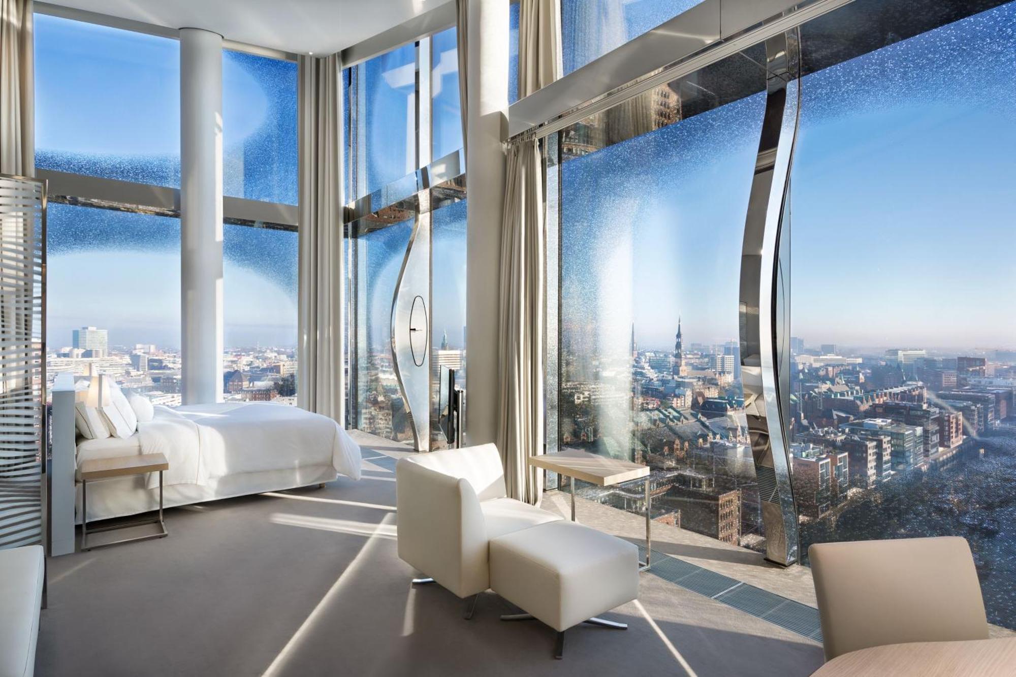 The Westin Hamburg Elbphilharmonie Hotell Eksteriør bilde The photo shows a modern hotel room with a stunning, panoramic view through large, uniquely designed windows. The room features a sleek, minimalist aesthetic with light-colored furnishings, including a white bed and a couple of chairs. The walls are 